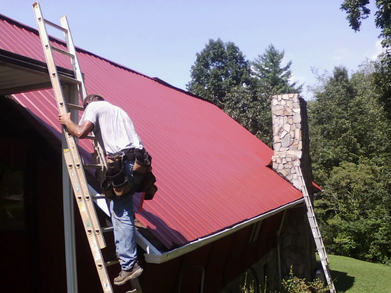 Roofer on a ladder beside a red metal roof and stone chimney — Soffit & Fascia Repair by Roofing Brothers in Batesburg